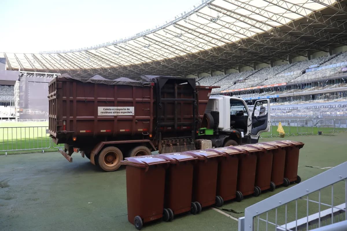 Caminhão de coleta ao lado de uma fileira de contentores marrons, na beira do gramado de um estádio.