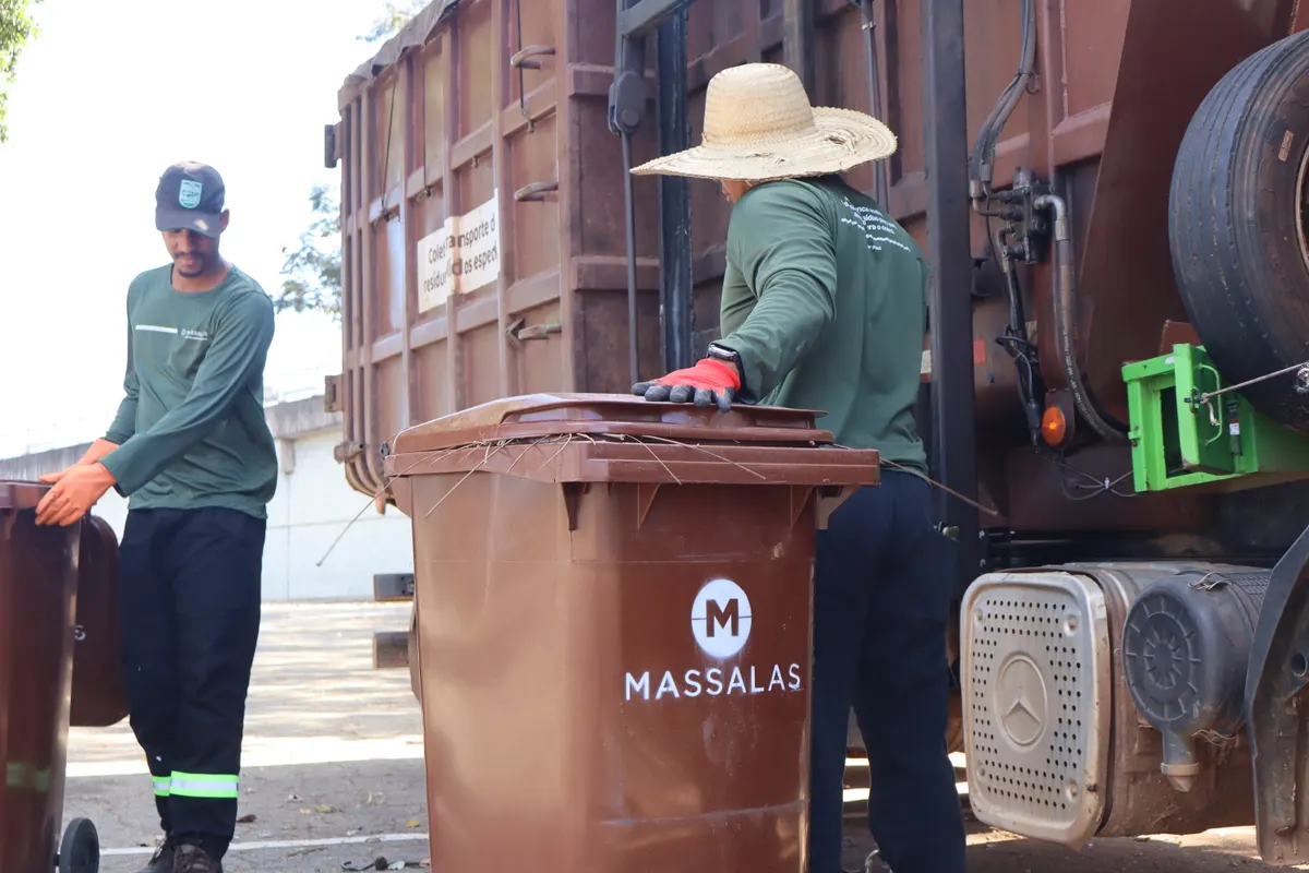 Dois operadores encaixando um contentor marrom no elevador hidráulico do caminhão de coleta.
