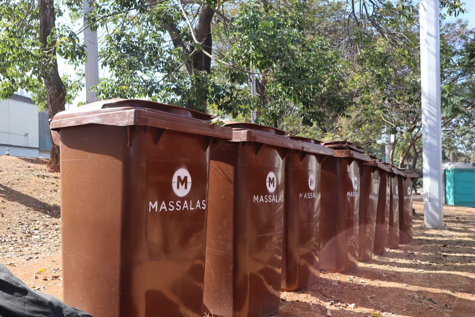 Fileira de contentores marrons com a marca da Massalas, alinhados sob as árvores do pátio de um cliente.