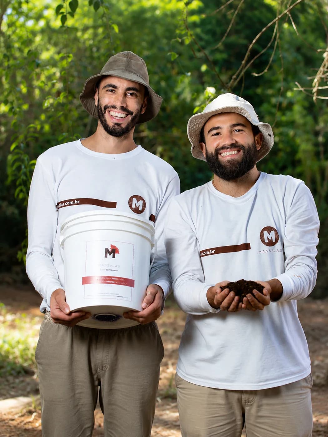 Guilherme Oliveira e Filipe Maciel, fundadores da Massalas, de chapéu e camiseta da marca: um segura um balde de composto e o outro mostra o composto pronto nas mãos.