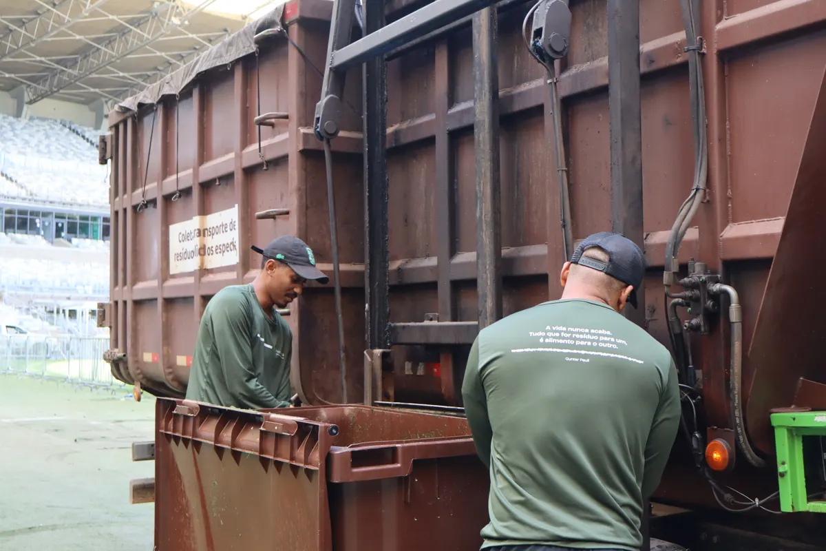 Dois operadores de uniforme descarregando um contentor marrom da traseira do caminhão de coleta.