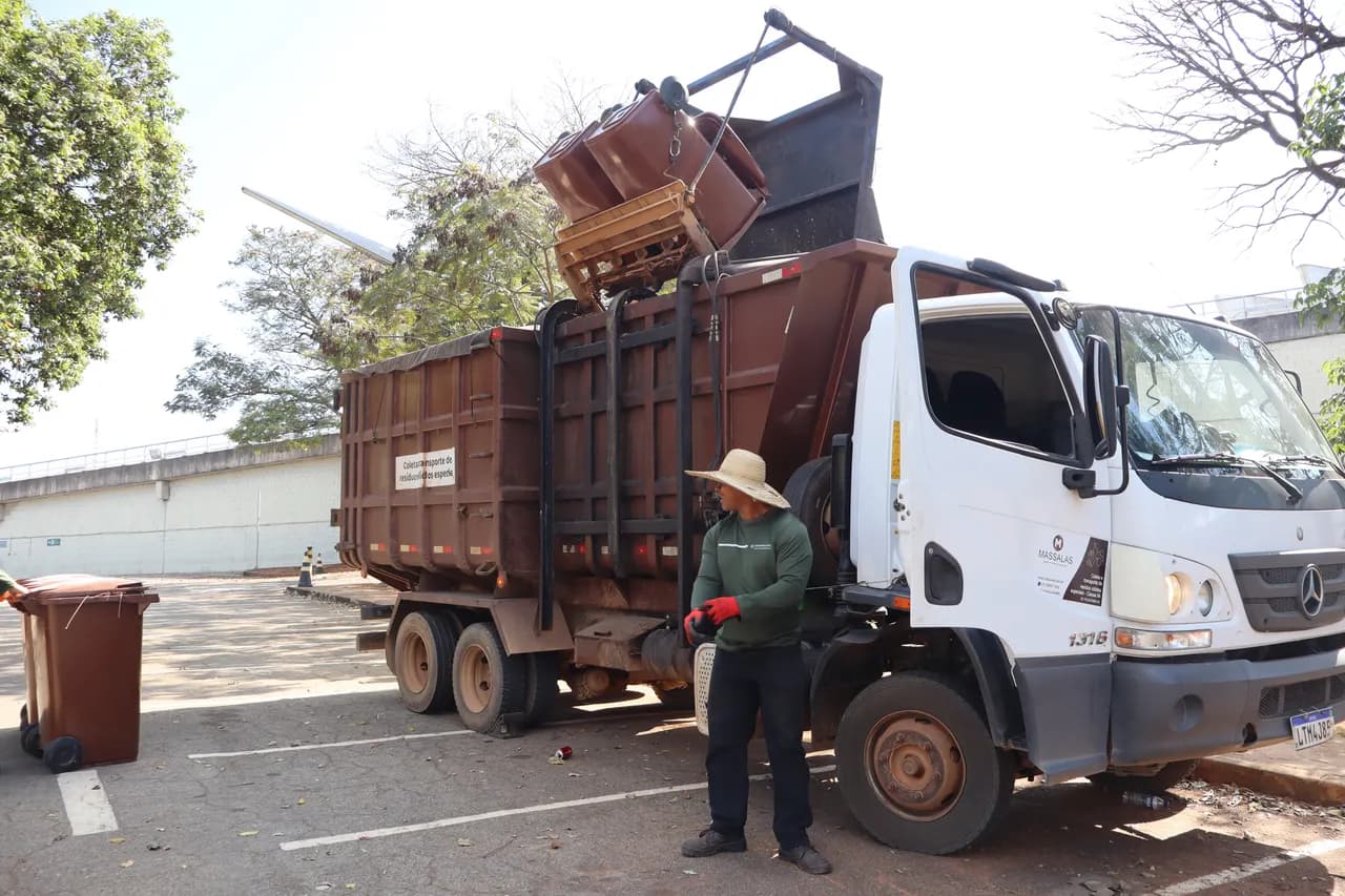 Caminhão de coleta com um contentor marrom erguido no elevador hidráulico e o operador ao lado, num estacionamento arborizado.