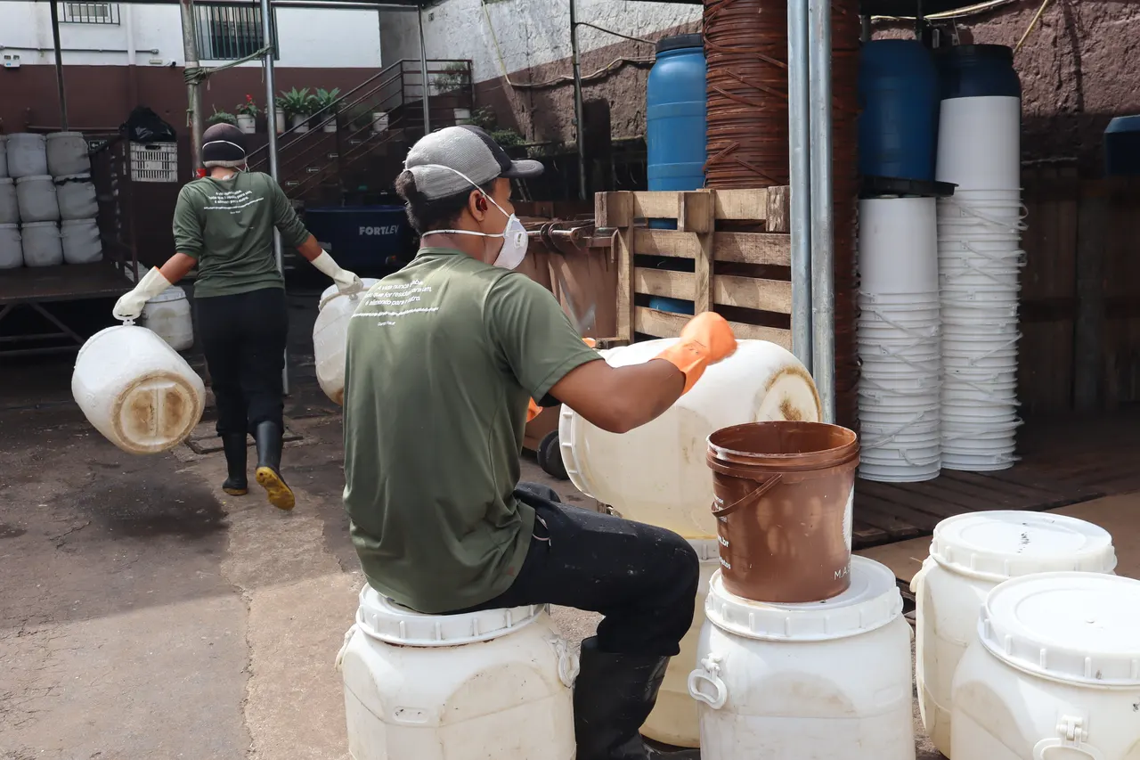 Dois operadores de máscara e luvas manejando bombonas brancas no pátio da unidade, com bombonas azuis e brancas empilhadas ao fundo.