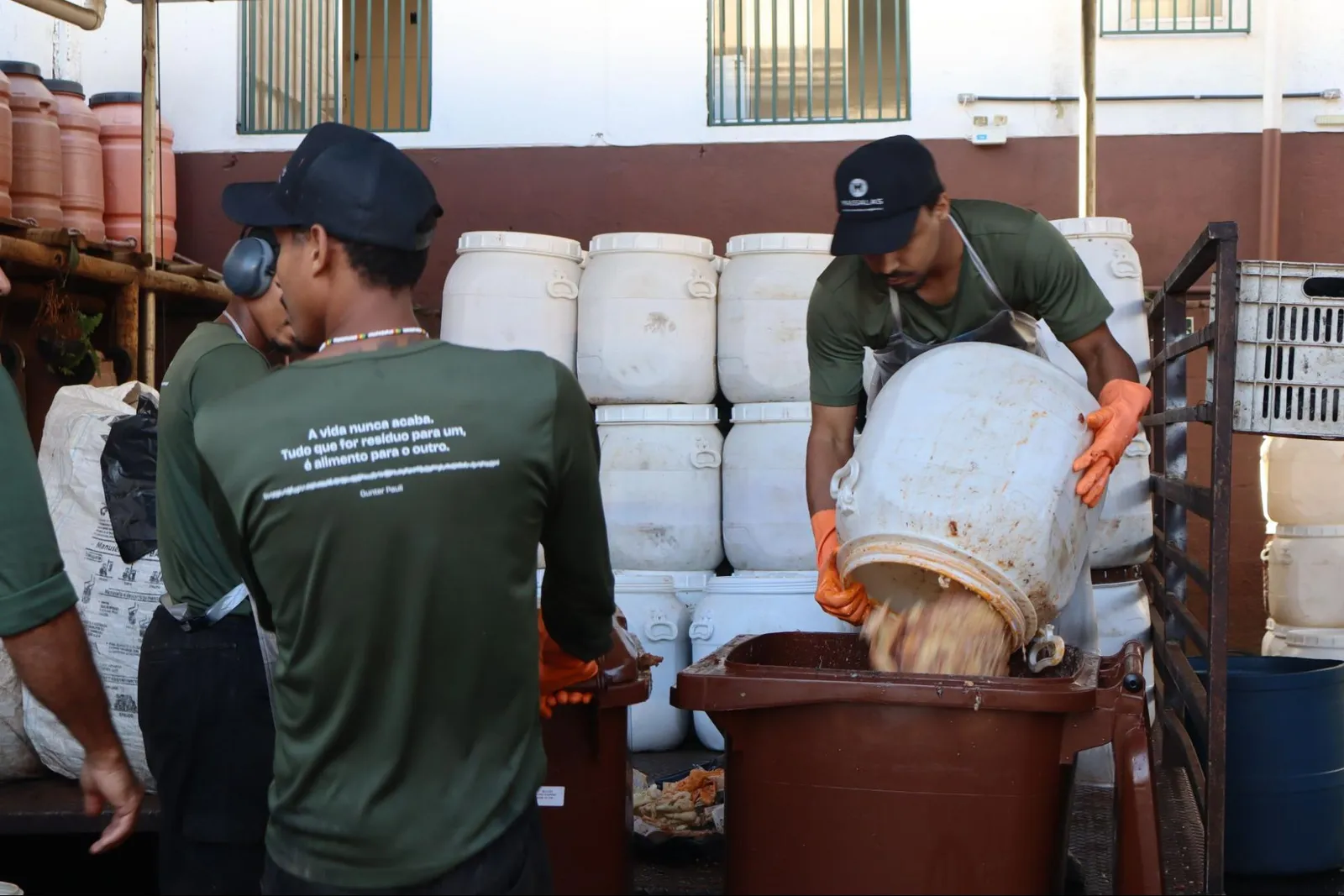 Operador de boné e luvas laranja virando uma bombona branca num contentor marrom, com a equipe e as bombonas empilhadas ao fundo.