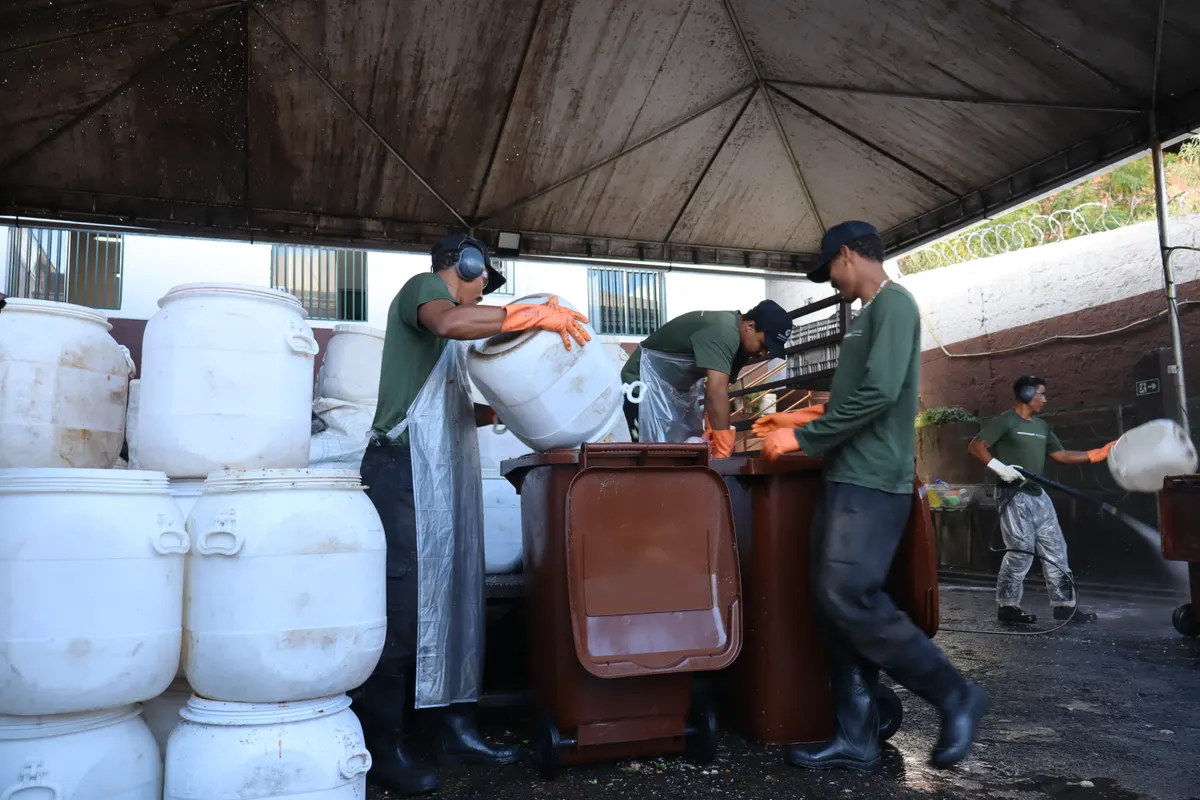 Operadores de luva esvaziando bombonas brancas em contentores sob a cobertura da unidade de triagem.